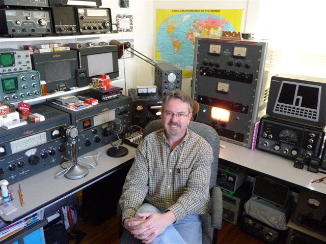 Clark sitting in between two tables of vintage equipment. Globe King, Hallicrafters, Drake and Heathkit equipment