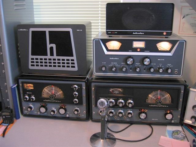 Old Hallicrafters station with HT32 transmitter and SX-115 receiver and transmitter and an additional older model receiver. Speaker with bit H sitting on top of the receiver and sitting on top of the HT32.  I see a SX-122 receiver sitting on top of the HT-32 transmitter with a Hallicrafters speaker sitting on top of that.