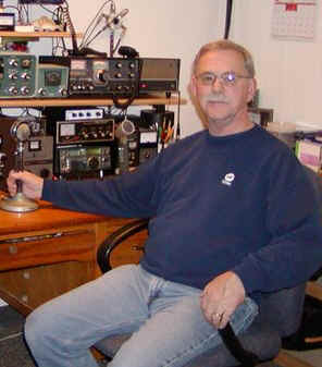 Karl sitting at his desk with right-hand on a D-104 microphone. On the dexk is a Collins 75A4 or similar receiver, what looks like a Johnson Viking II and some newer SSB equipment. On a shelf above the desk I see a Drake 2-B, Heathkit S-line transceiver with external VFO and a Swan 350 transceiver. On a shelf above that I see an old Hallicrafters receiver and on a shelf above that a couple of speakers a call sign plaque and other electronic gear. 