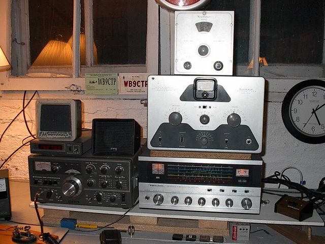 Down in Ron's basement we have a Kenwood TS-520S. Sitting on top of the Kenwood is a Moitorola Motrac speaker, and digital display. To the right of the Kenwood is the vintage AM stack consisting of (from top to bottom) Heathkit VF-1 VFO, DX-20, and Realistic DX-160 receiver. On the operating desk is Ron's straight key for slow speed CW. All radios are sitting on a shelf above the operating desk. Under the receiver we see 4 crystals for the DX-20.