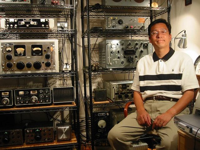 Masa sitting  next to two racks of radios. Each rack has four or five shelves loaded with cool equipment. You will have to ask Masa what it all is.