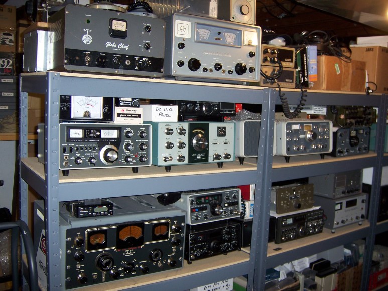 Three level storage rack about three units wide containing Globe chief, Hammarlund receiver, old Yaesu transceiver, HW101, another transceiver, Hallicrafters Sky Buddy, and four or five more transceivers and I can't see what's on the lower level